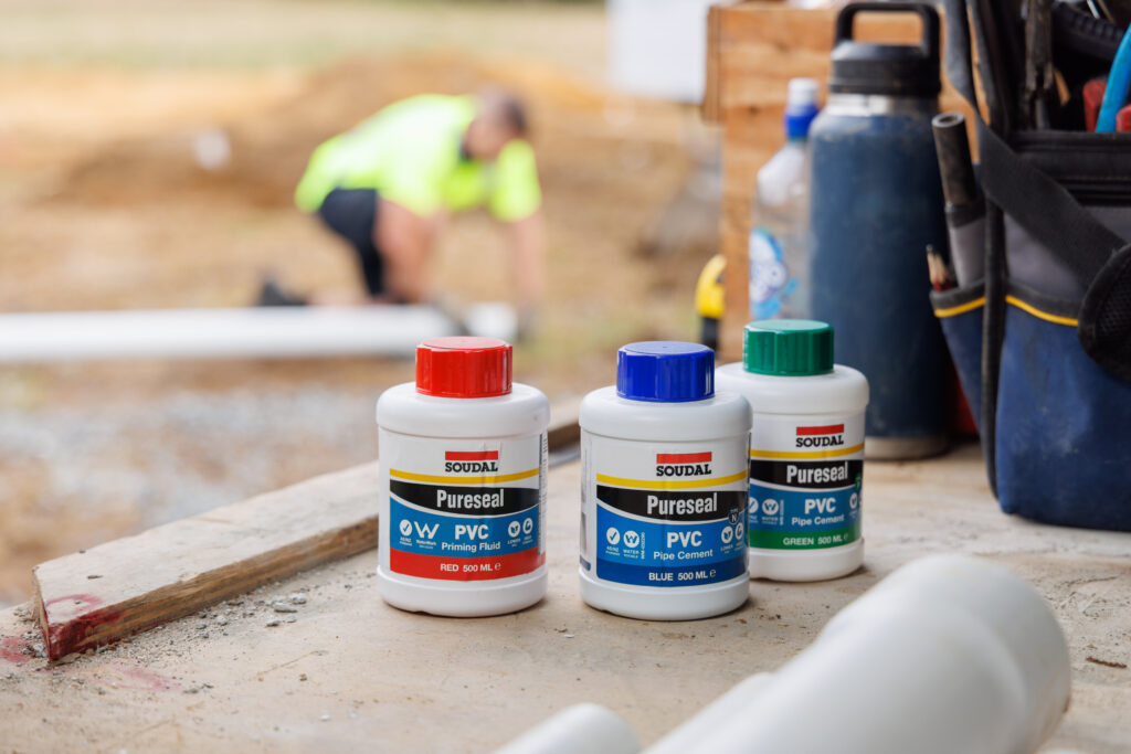 Soudal Products This image displays three bottles of Soudal Pureseal products used in PVC pipe installations: Red Cap: PVC Priming Fluid Blue Cap: PVC Pipe Cement Green Cap: PVC Pipe Cement A technician is visible in the blurred background, working on piping, which aligns with the products’ intended use
