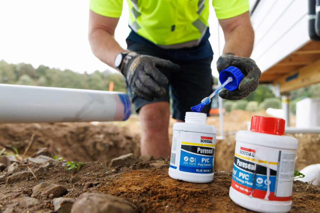 Soudal Products This image shows a tradesperson in high-vis workwear and gloves applying Soudal Pureseal PVC pipe cement outdoors on a job site. Two Pureseal containers are in focus—one with a blue lid (PVC Pipe Cement) and another with a red lid (PVC Pipe Priming Fluid). The scene highlights professional-grade use of the product in real-world plumbing applications.